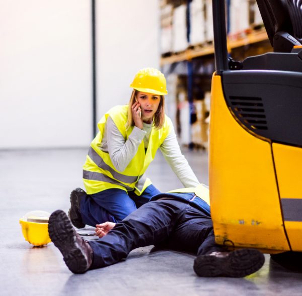 An accident in a warehouse. Woman with smartphone and her colleague lying on the floor next to a forklift.